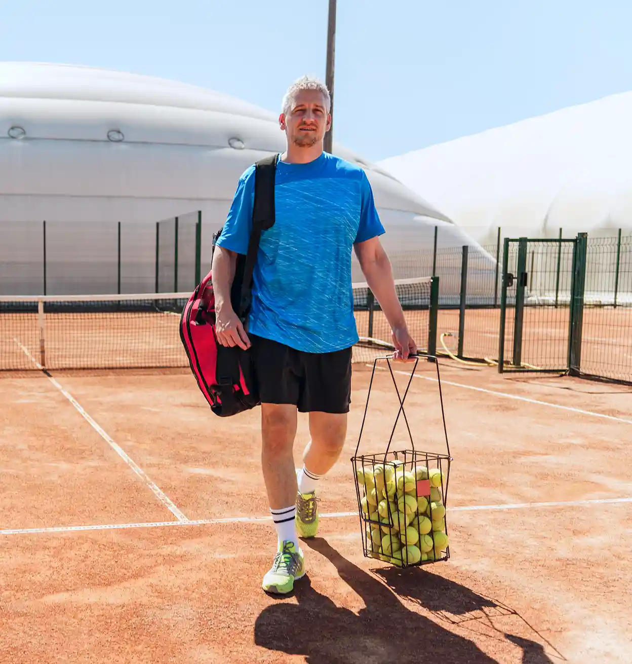 a man on a tennis court carrying a bag and a basket of balls